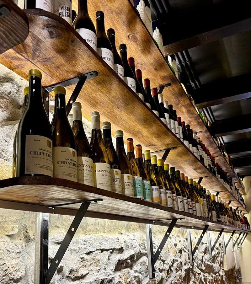 Filas de botellas de vino en estantes rústicos de madera contra una pared de piedra en una bodega.