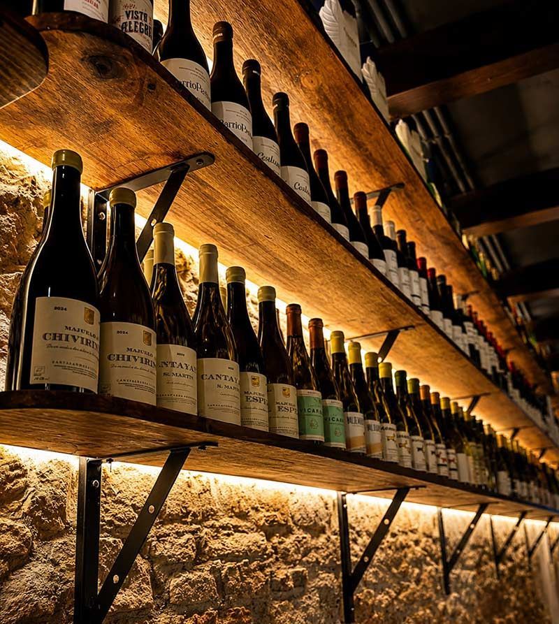 Botellas de vino iluminadas con una luz cálida, expuestas en estantes de madera en una bodega rústica.