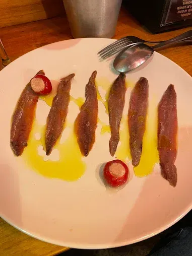 Anchoas servidas en un plato blanco con aceite de oliva, dos pimientos rojos con queso, tenedor y cuchara.
