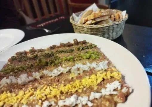 Primer plano de un plato en capas sobre un plato blanco con una canasta de pan en el fondo, sobre una mesa oscura.