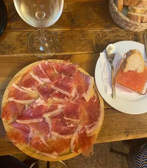 Jamón curado en lonchas en bandeja de madera con pan, mantequilla y copa de vino sobre una mesa de madera.