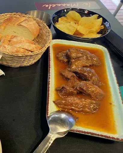 Plato de carne guisada con salsa, cesta de pan y bol de patatas fritas sobre una mesa.