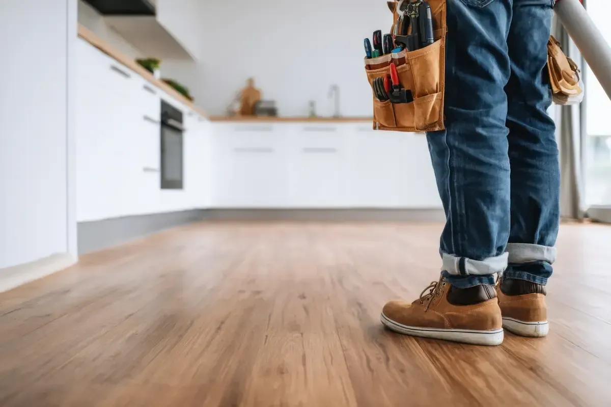 Persona de pie en una cocina luminosa, vestida con vaqueros, botas de trabajo y un cinturón de herramientas con herramientas.