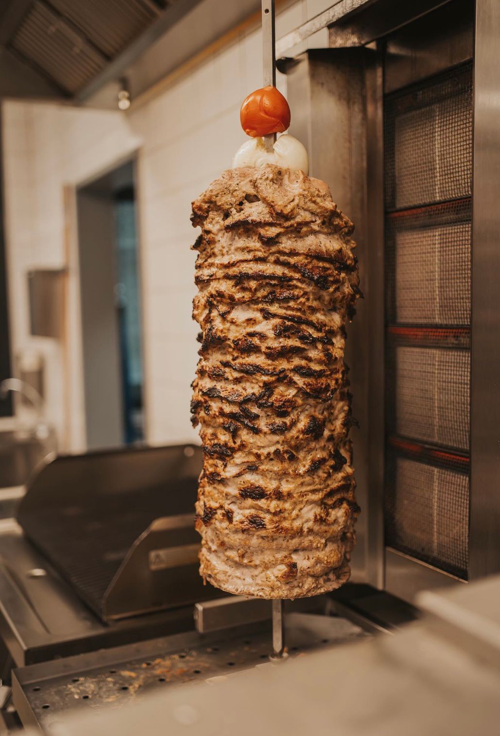 Vertikaler Drehspieß mit geschichtetem Fleisch, der sich in einer Küche dreht und mit einer Tomate garniert ist.
