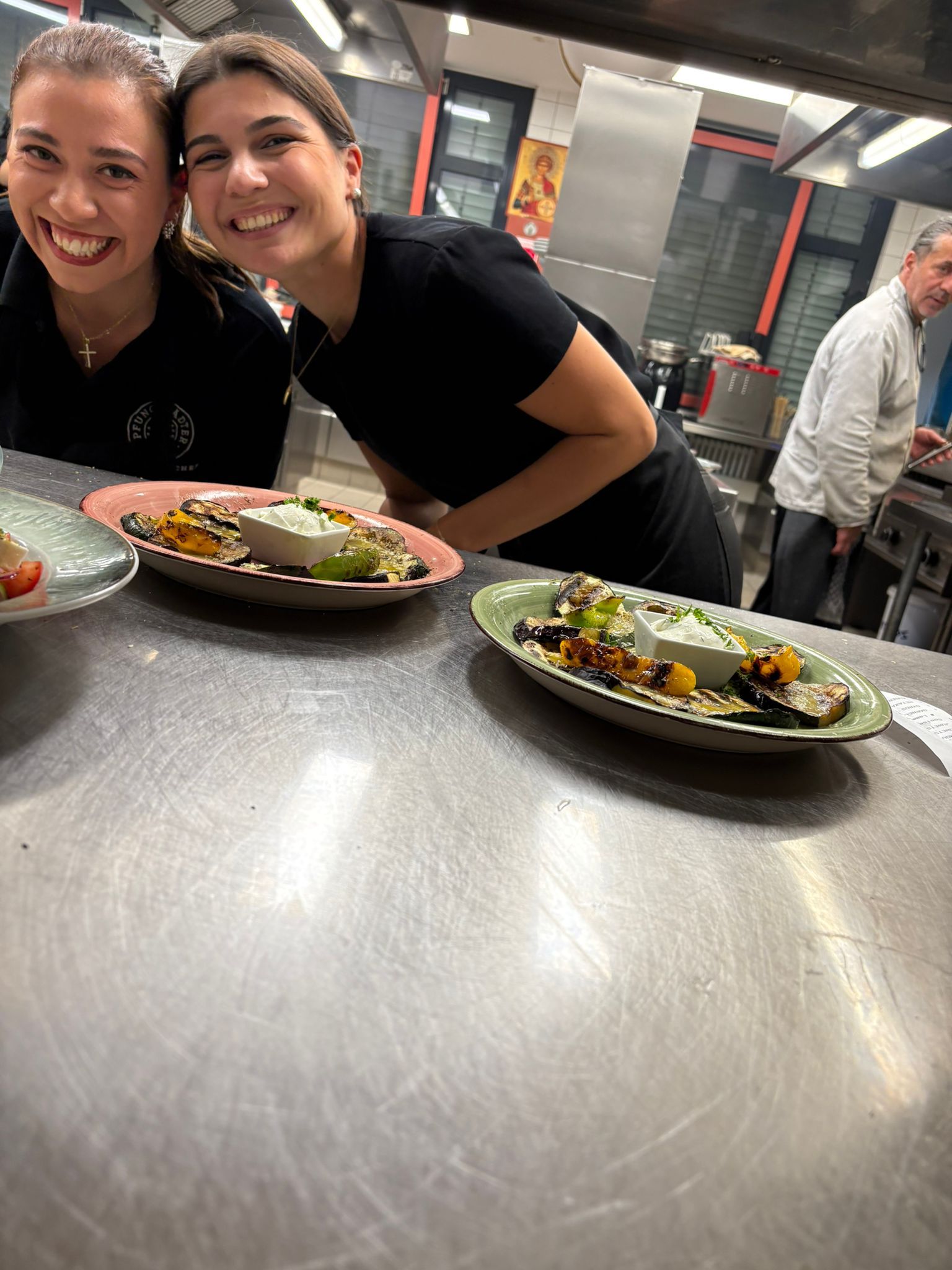 Zwei lächelnde Freunde beugen sich mit auf Tellern servierten Speisen über eine Theke in einer Restaurantküche.