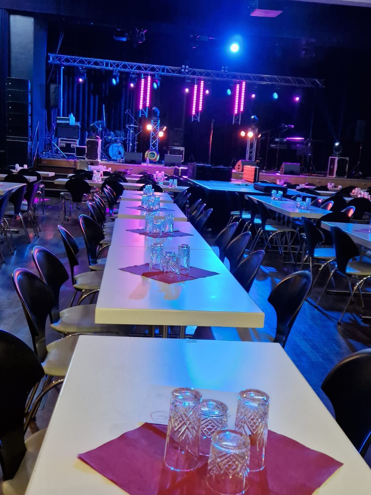 Festtafeln sind für eine Veranstaltung in einer schwach beleuchteten Stadthalle mit blau-violetten Bühnenlichtern gedeckt.