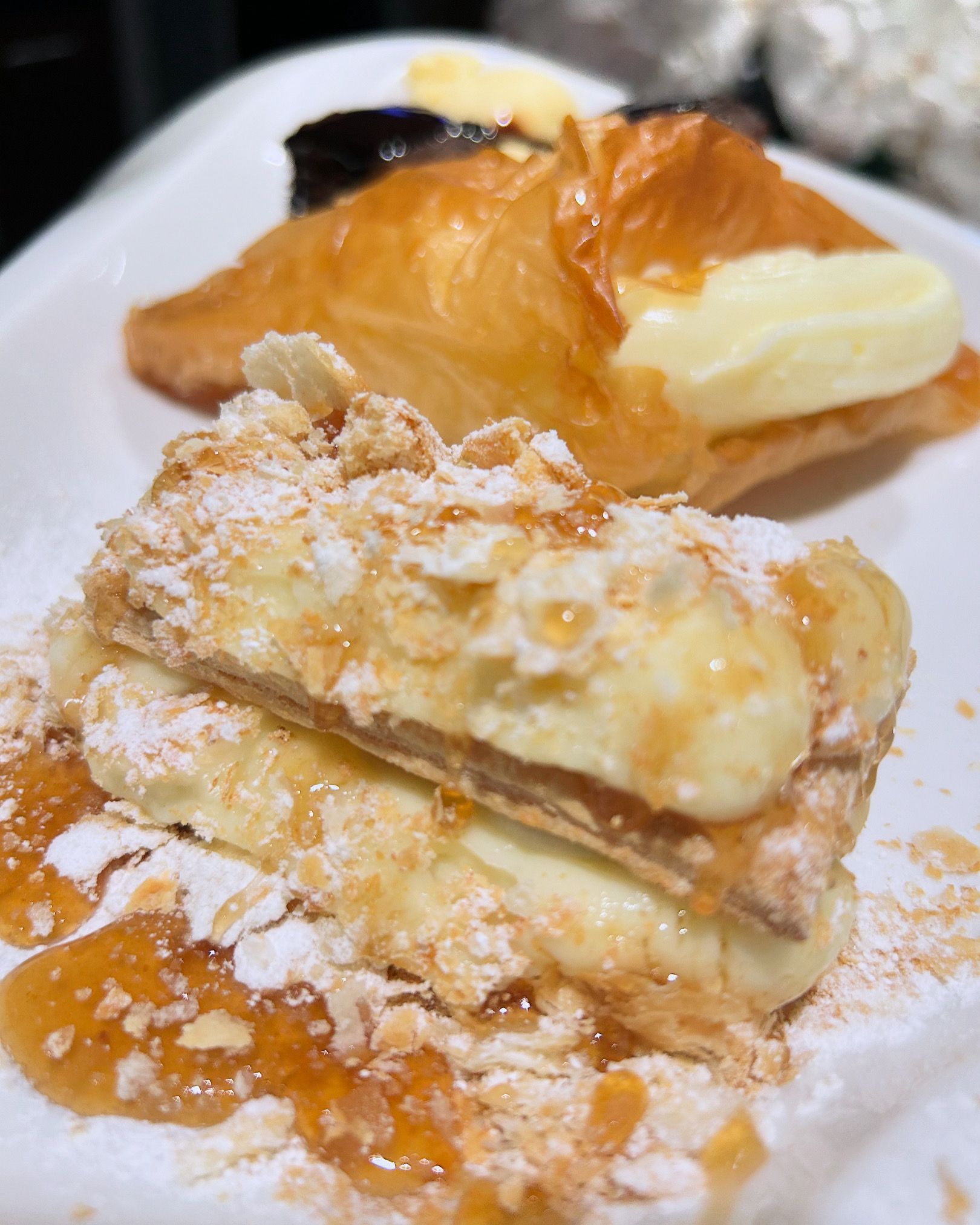 Gebäckdessert mit Puderzucker und Karamellsauce auf einem weißen Teller, dahinter ein Croissant und Butter.