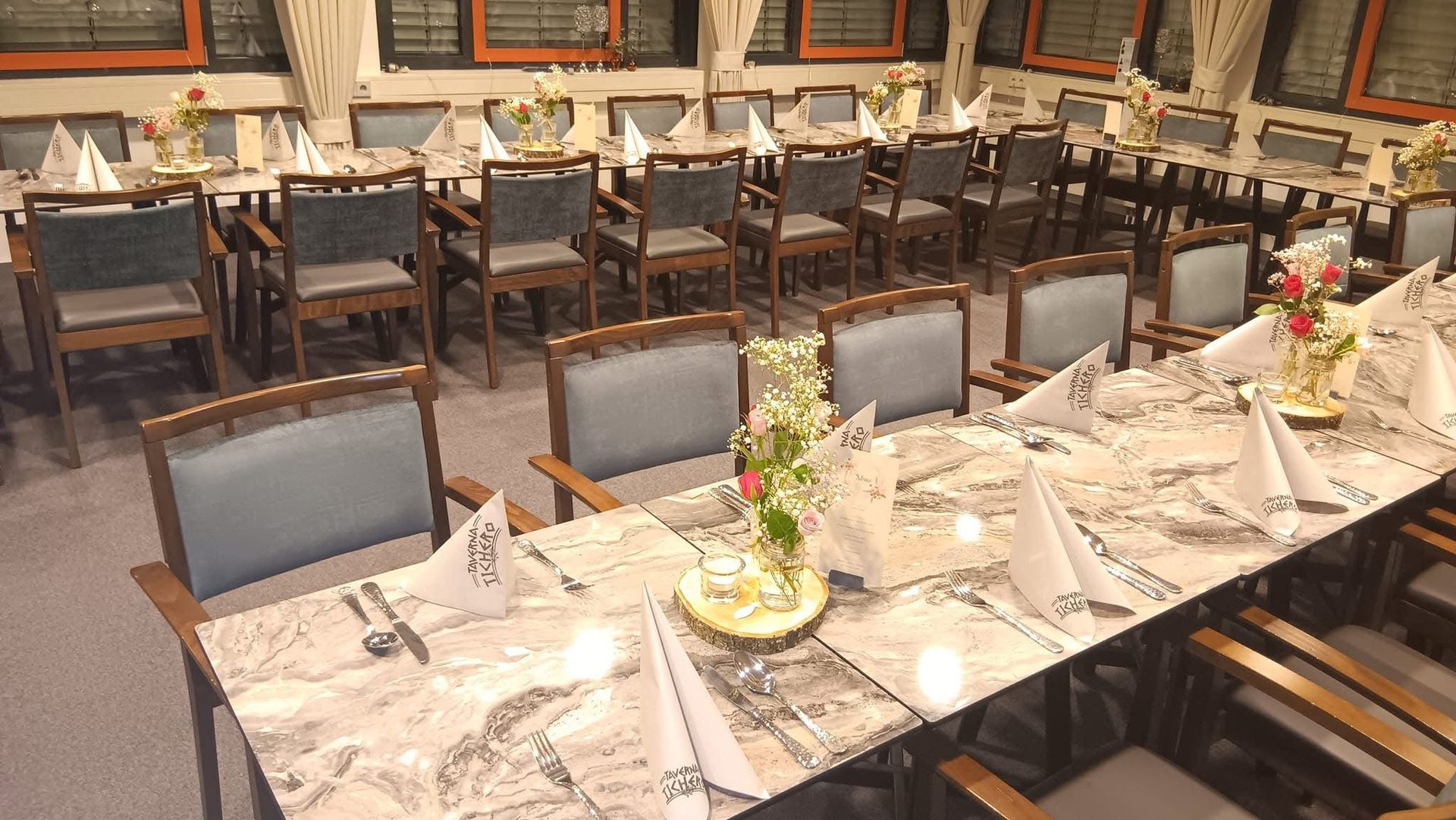 Elegantes Esszimmer mit gedeckten Tischen, Stühlen und mit Kerzen beleuchteten Blumenarrangements auf Marmortischplatten.