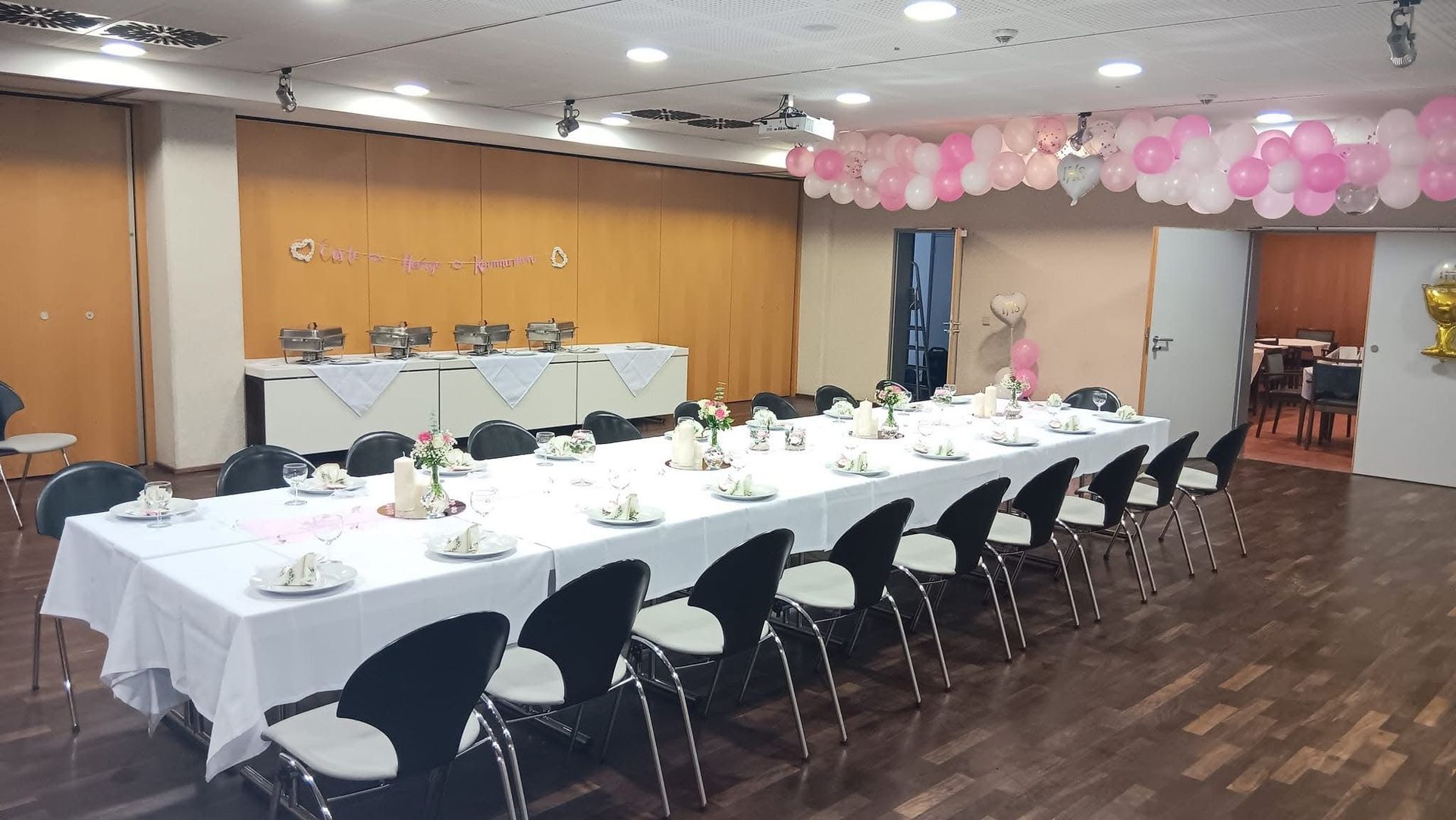 Festsaal mit langen, weiß gedeckten Tischen, schwarzen Stühlen und rosa Ballongirlanden an der Wand.