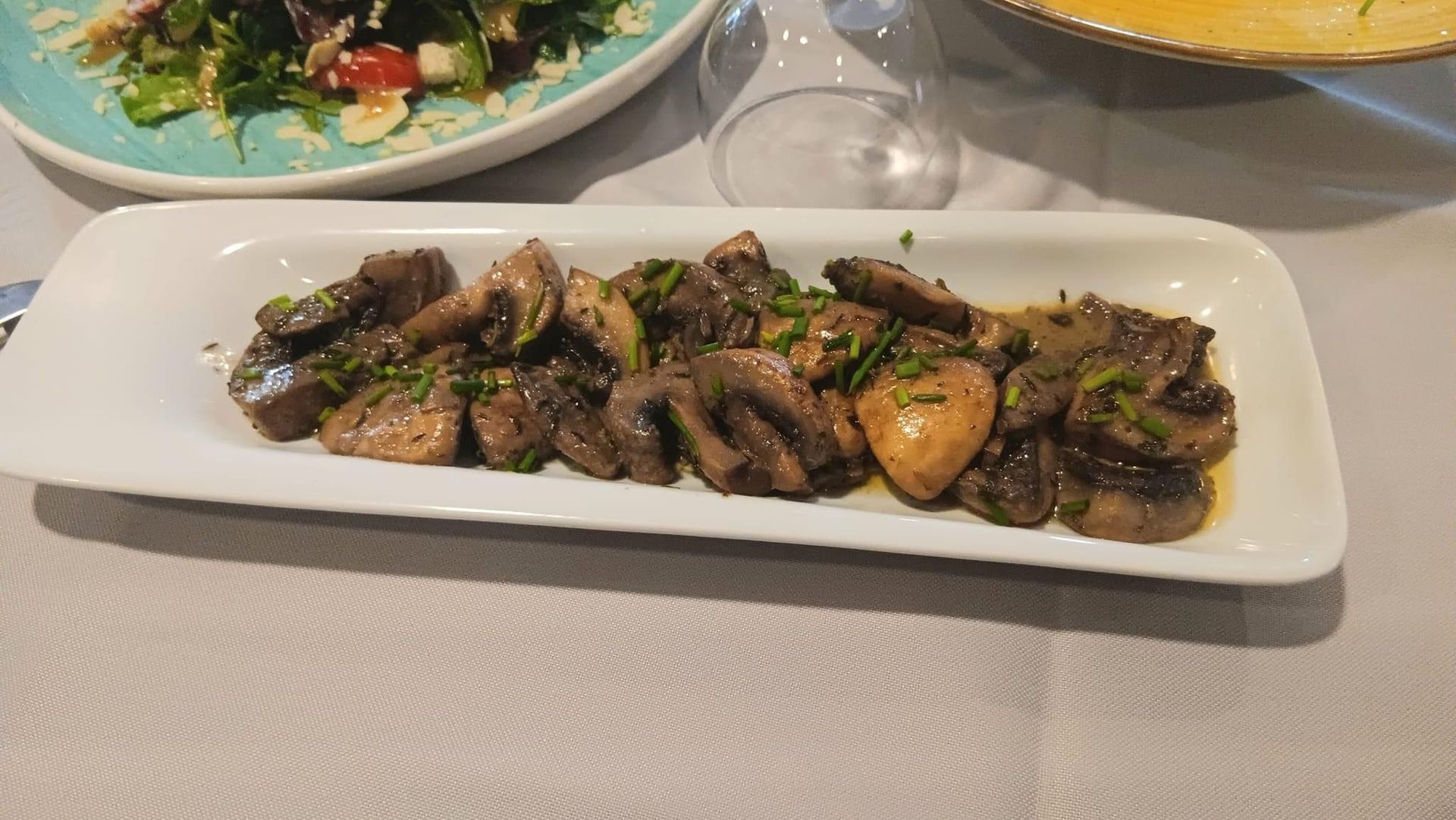 Gegrillte Champignons und Fleisch auf einem weißen, rechteckigen Teller angerichtet, im Hintergrund Salate.