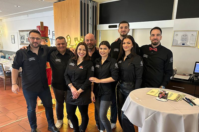 Eine Gruppe von Mitarbeitern in schwarzen Uniformen posiert drinnen neben einem runden Tisch mit Dessert und Getränken.