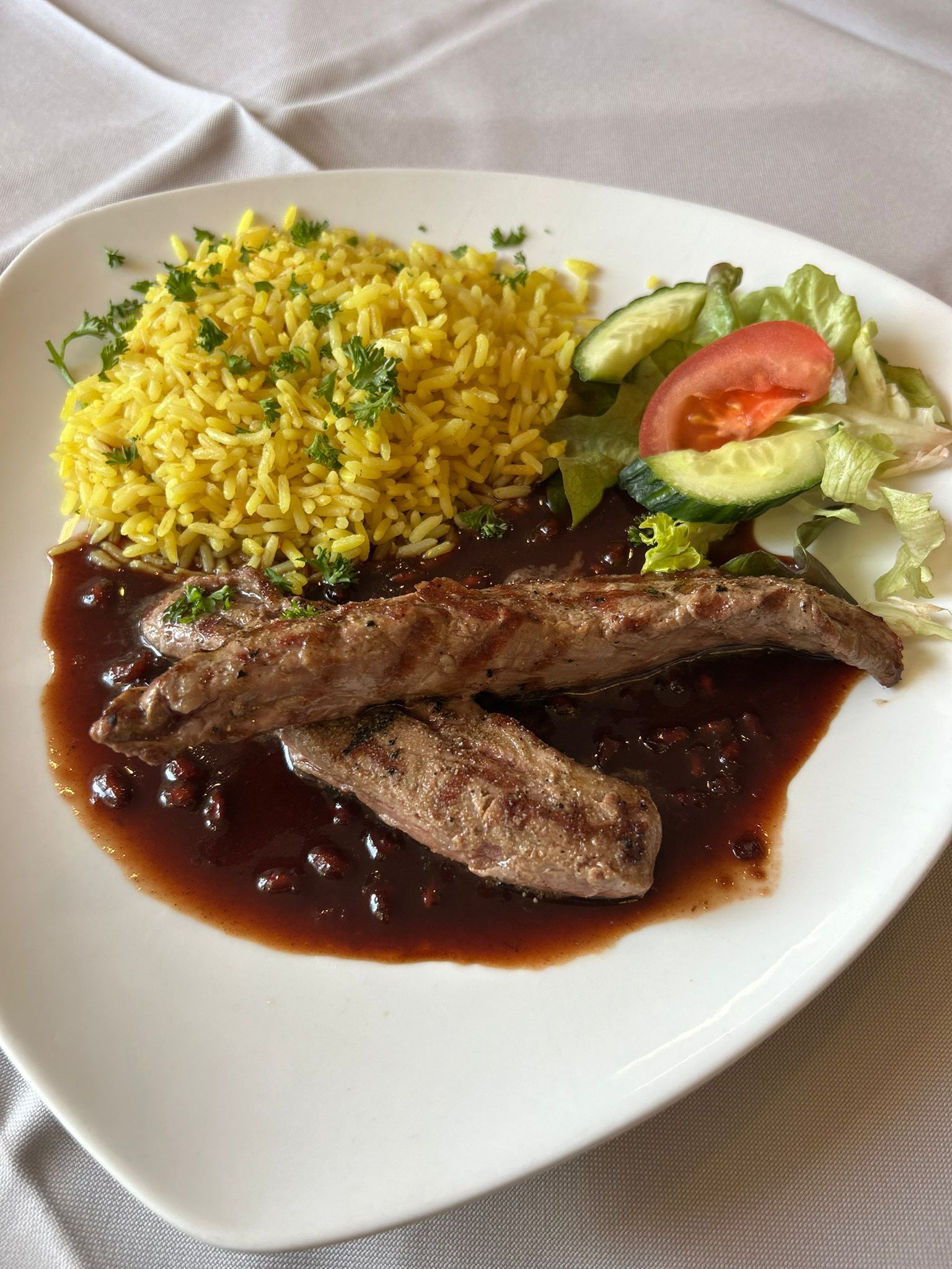 Teller mit gelbem Reis, gegrilltem Fleisch in dunkler Soße und einem kleinen Salat mit Gurke und Tomate.
