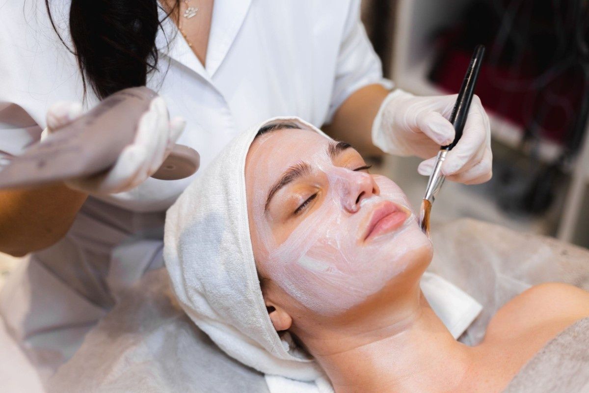 Mujer recibiendo tratamiento facial; esteticista aplicando mascarilla con cepillo.