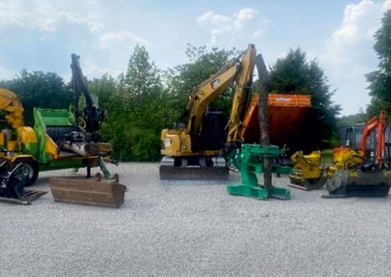 Draußen auf einem Kiesplatz aufgereiht stehen Baumaschinen: Bagger, Häcksler, orangefarbener Muldenkipper und andere Maschinen.