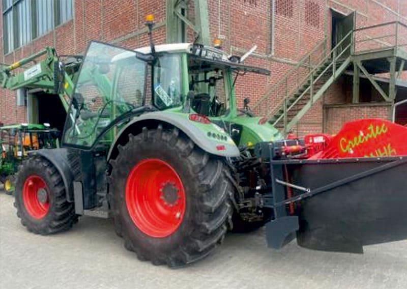 Grüner Traktor mit roten Rädern und Mähaufsatz, neben einem Backsteingebäude geparkt.