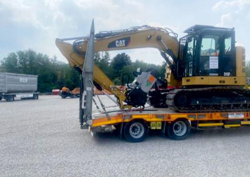 Gelber Caterpillar-Bagger auf einem Anhänger, bereit zum Transport, geparkt auf einem Schotterplatz.