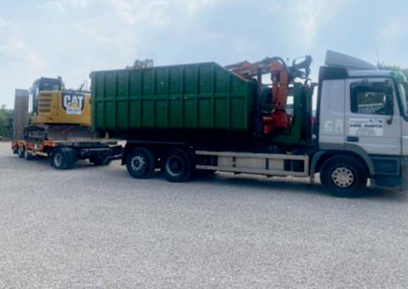 LKW, der Baumaschinen transportiert: einen Bagger auf einem Anhänger, einen großen grünen Müllcontainer und einen Kran.