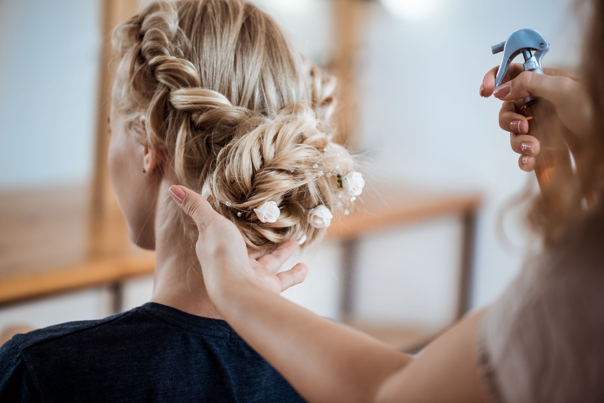 Eine Person stylt blondes Haar zu einer Hochsteckfrisur, während eine andere es im Salon mit Haarspray fixiert.