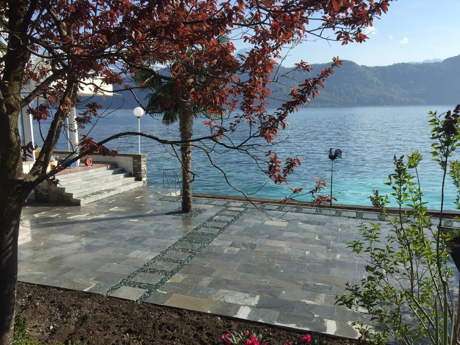 Terrasse am See mit rotblättrigem Baum, Steinstufen und türkisfarbenem Wasser unter blauem Himmel.