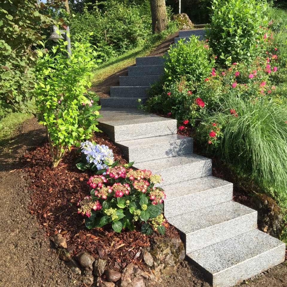 Steinstufen führen einen Hügel hinauf, der von blühenden Büschen und grünem Laub gesäumt ist.