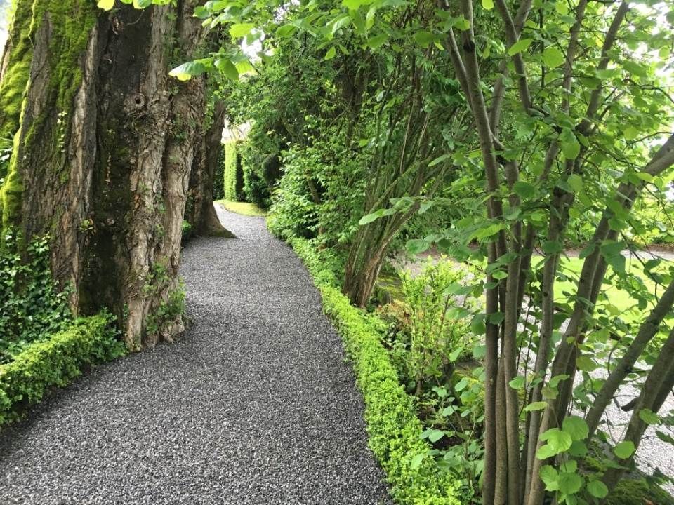 Ein Kiesweg führt durch einen Garten, gesäumt von geschnittenen grünen Hecken und Bäumen.