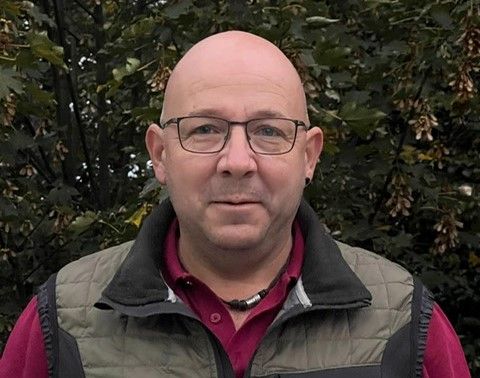 Glatzköpfiger Mann mit Brille, der eine Weste und ein rotes Hemd trägt, lächelt vor einer Laubwand.