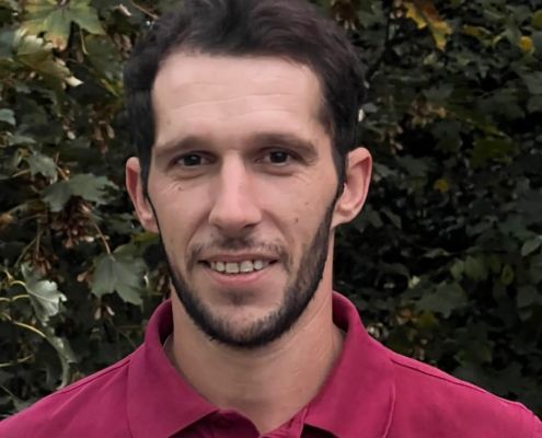 Mann mit dunklem Haar und Bart, der ein rotes Hemd trägt, lächelt im Freien vor grünem Laubwerk.