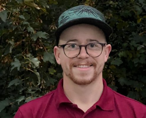 Ein Mann mit Brille und Baseballkappe lächelt vor einem grünen Hintergrund und trägt ein weinrotes Poloshirt.