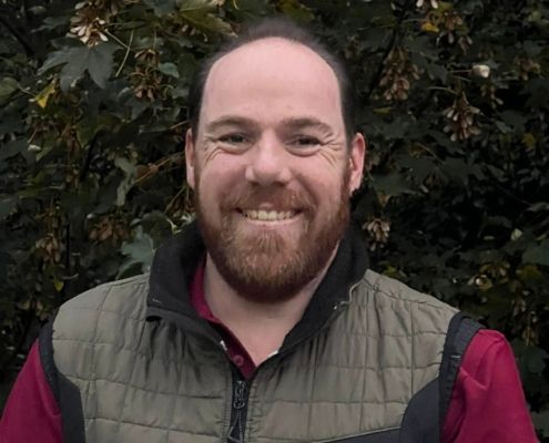 Ein lächelnder Mann mit Bart, der eine grüne Weste über einem roten Hemd trägt, steht vor dichtem Laubwerk.