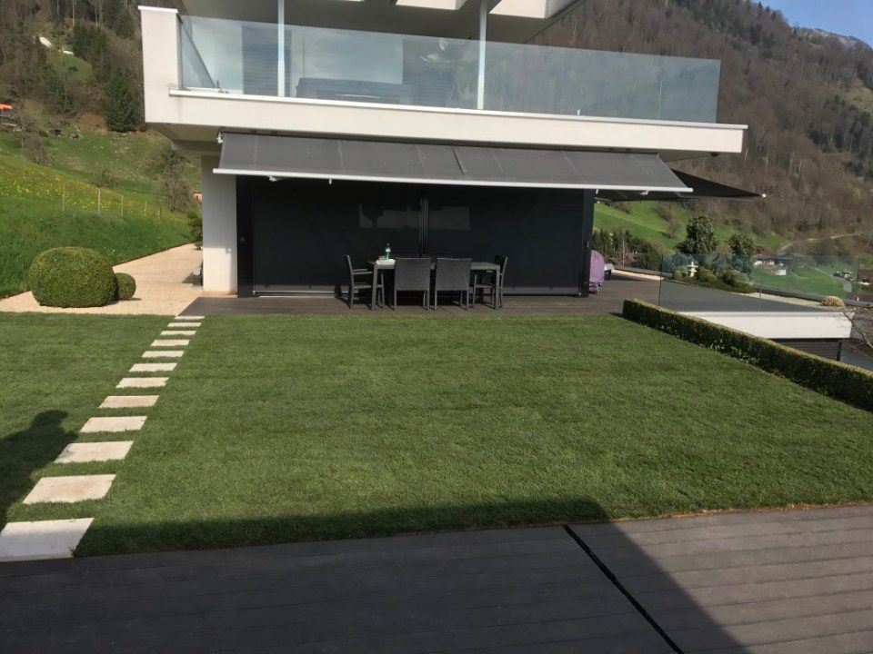 Modernes Haus mit dunkler Markise über der Terrasse, grünem Rasen, Steinweg und Bergkulisse.