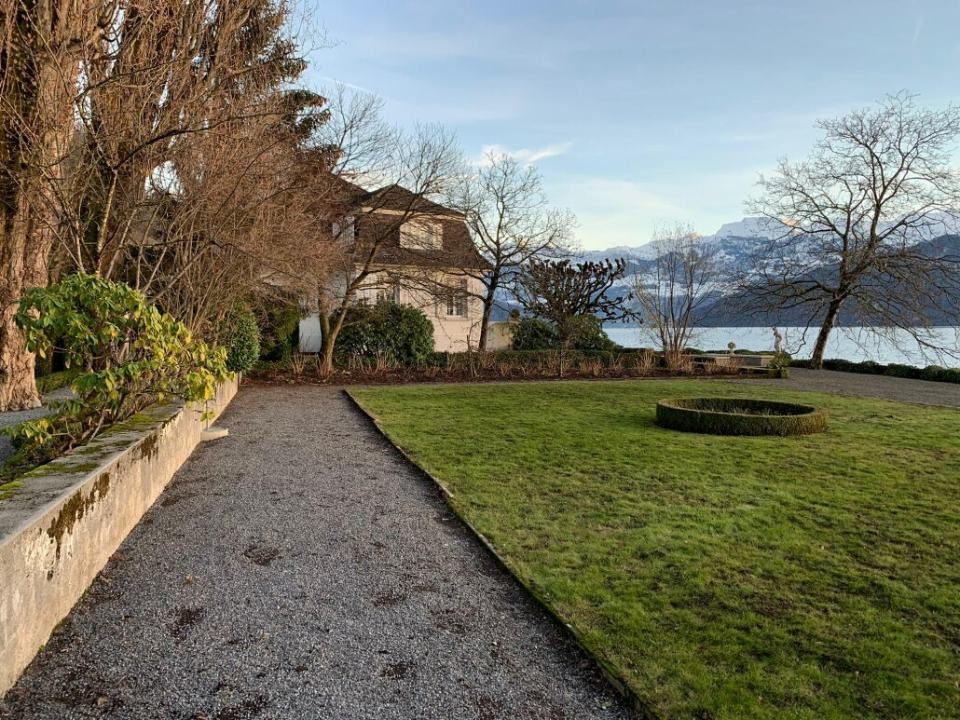 Ein Kiesweg führt durch einen Garten zu einem Haus, einem See und schneebedeckten Bergen unter einem teilweise bewölkten Himmel.