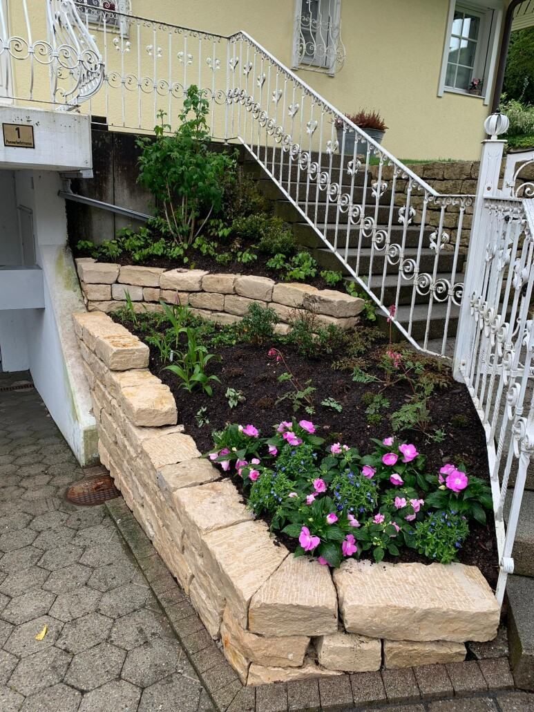 Ein von Steinmauern umgebenes Gartenbeet mit rosa Blumen und grünen Pflanzen neben einer Treppe.