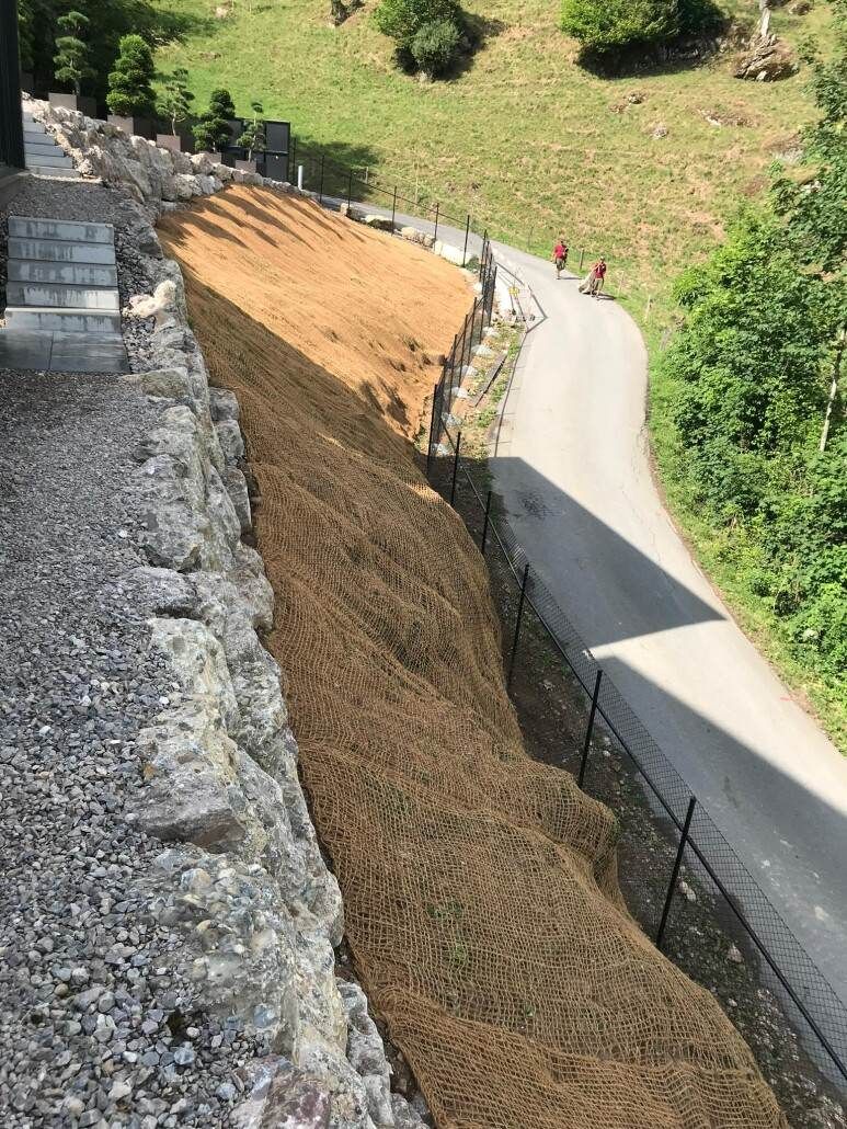 Ein mit braunem Netz bedeckter Straßenrandwall entlang einer kurvenreichen Strasse in einer grünen, hügeligen Gegend.