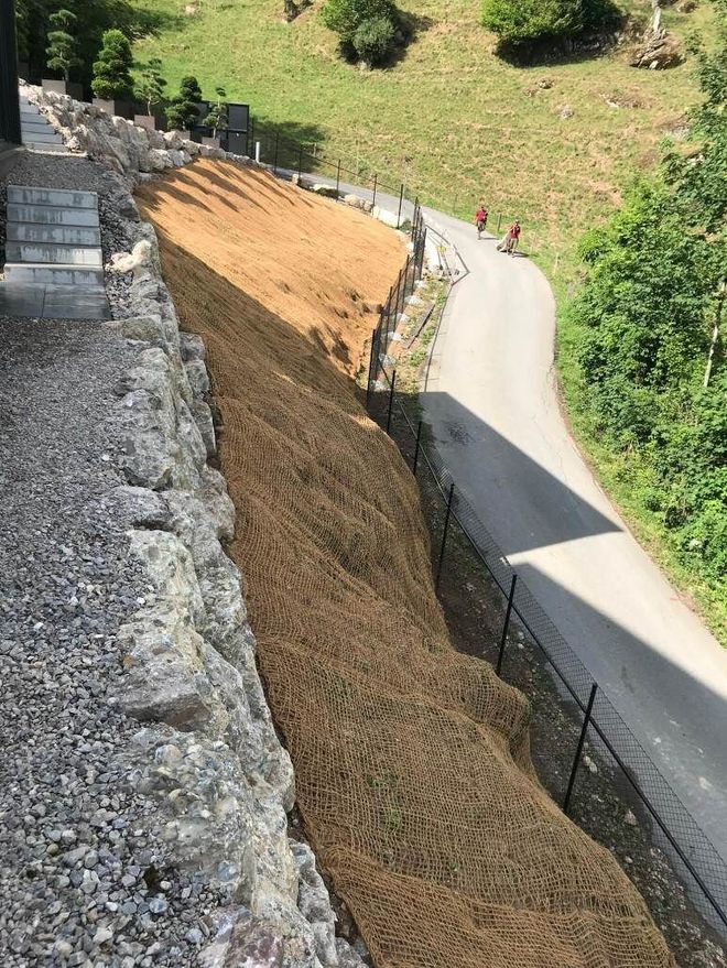 Ein mit braunem Netz bedeckter Straßenrandwall entlang einer kurvenreichen Strasse in einer grünen, hügeligen Gegend.