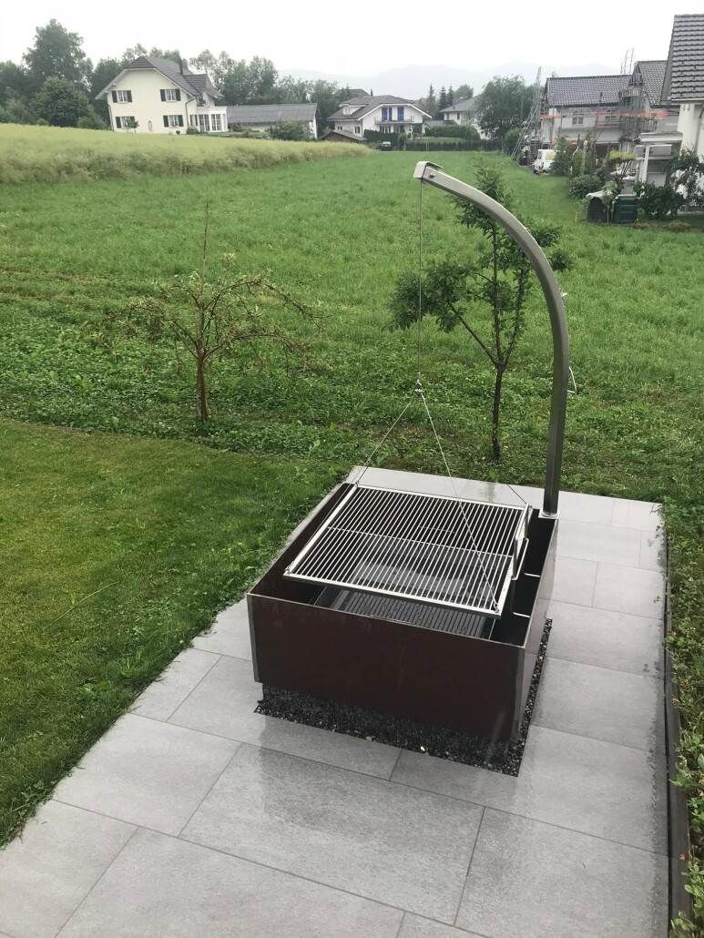 Grillplatz im Freien auf einer Steinterrasse mit Blick auf eine Wiese und Häuser im Hintergrund.