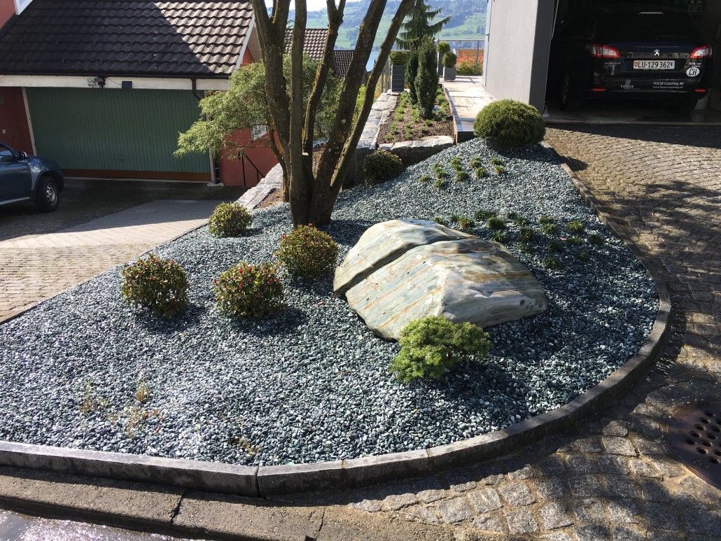 Ein angelegtes Gartenbeet mit blauem Kies, Findlingen, Sträuchern und einem Baum, neben einer Einfahrt.