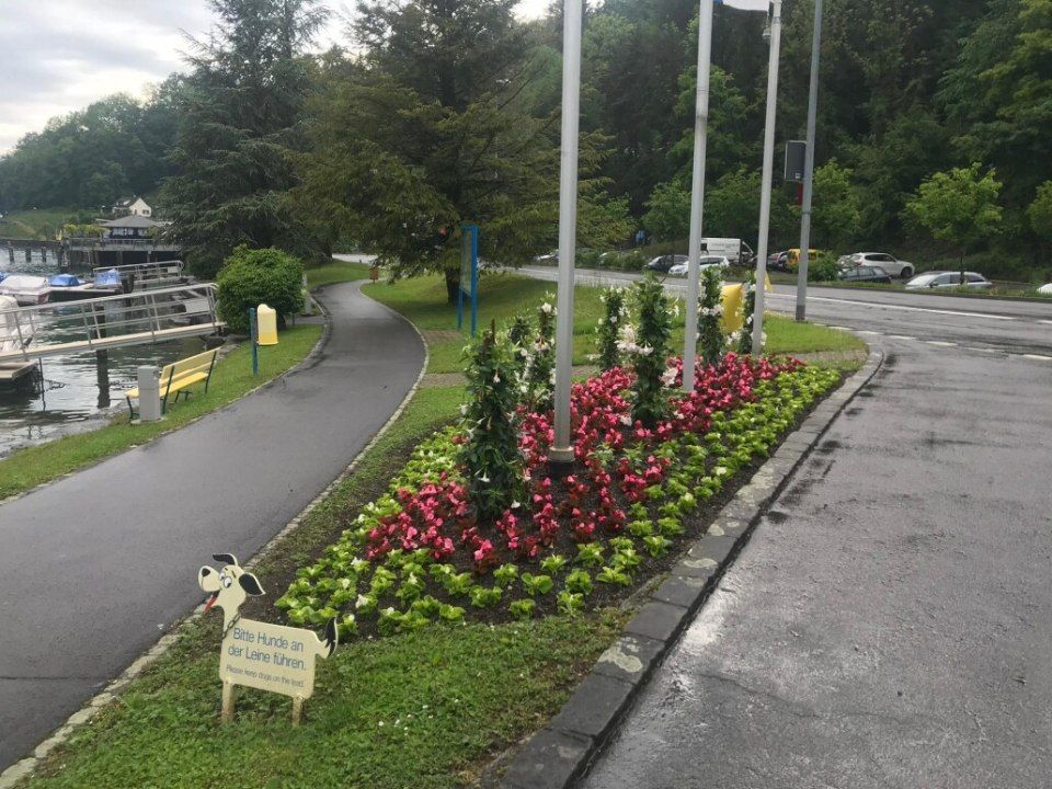 Weg und Blumenbeet neben einem See mit einer kleinen Ziegenstatue im Vordergrund.