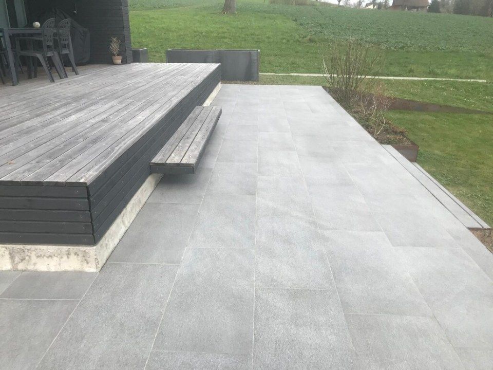 Eine grau geflieste Terrasse grenzt an eine erhöhte Holzterrasse mit eingebauter Bank. Im Hintergrund ist grünes Gras zu sehen.
