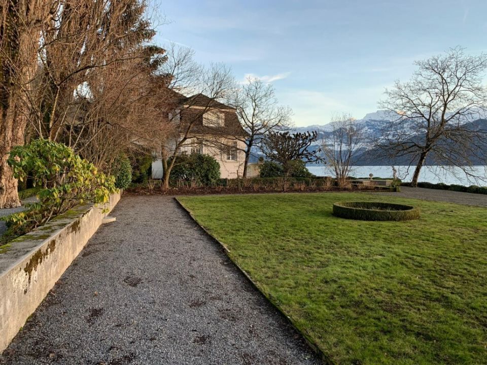 Ein Schotterweg führt zu einem weissen Haus an einem See, umgeben von Bäumen und einem grünen Rasen an einem sonnigen Tag.