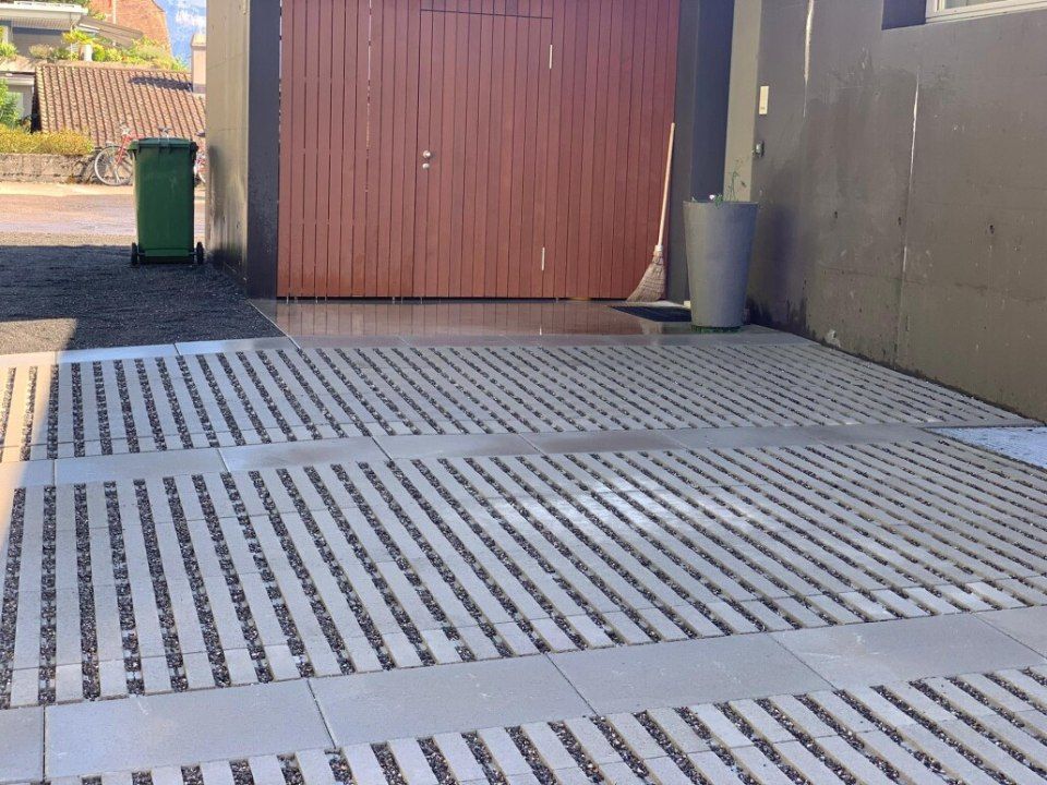 Braunes Holztor, betonierte Auffahrt mit Kiesabschnitten, grüne Mülltonne, grauer Container, Sonnenlicht.