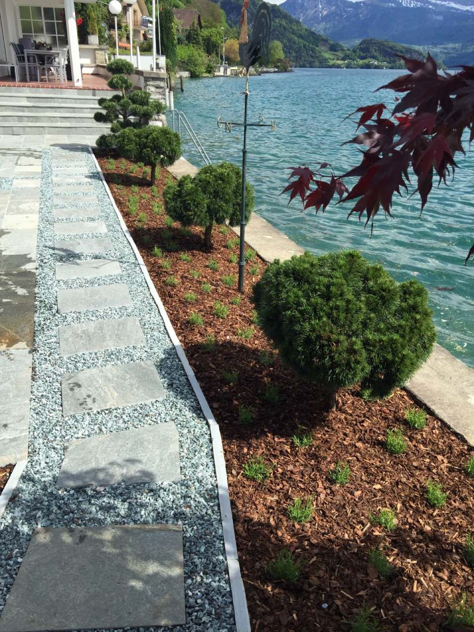 Garten am See mit Weg, kleinen Formgehölzen und Bergen im Hintergrund. Blaues Wasser und ein rotblättriger Baum.