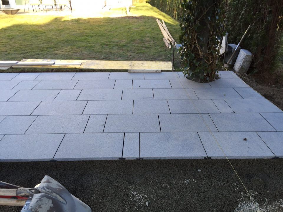 Eine Terrasse aus grauem Stein liegt in einem Garten, im Hintergrund sind Gras und Grünpflanzen zu sehen.