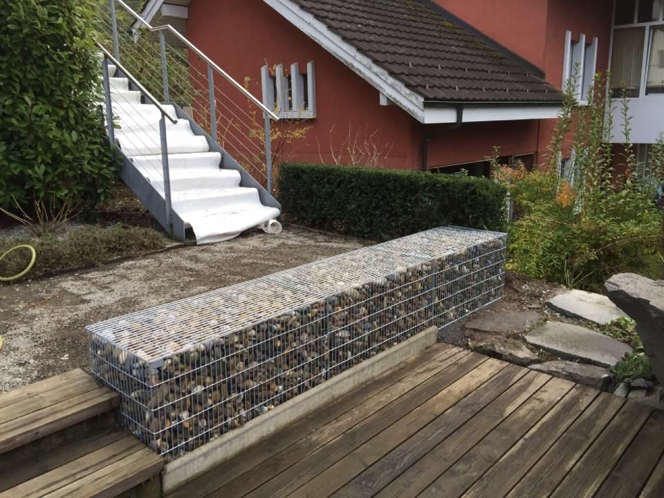 Gabionen-Stützmauer aus Drahtgeflecht, gefüllt mit Steinen, in der Nähe einer Holzterrasse und einer Aussentreppe.