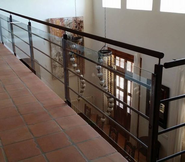 Vista interior de barandilla de balcón con barras metálicas horizontales, paneles de vidrio y pasamanos de madera, piso de baldosas de terracota.