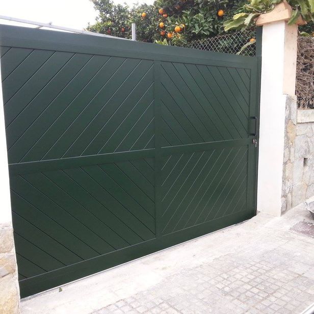 Portón metálico de color verde con diseño diagonal, frente a un muro de piedra y un naranjo.