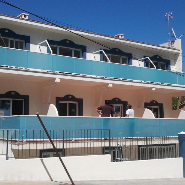 Edificio de dos pisos con detalles azules, dos hombres de pie en el balcón, día soleado.