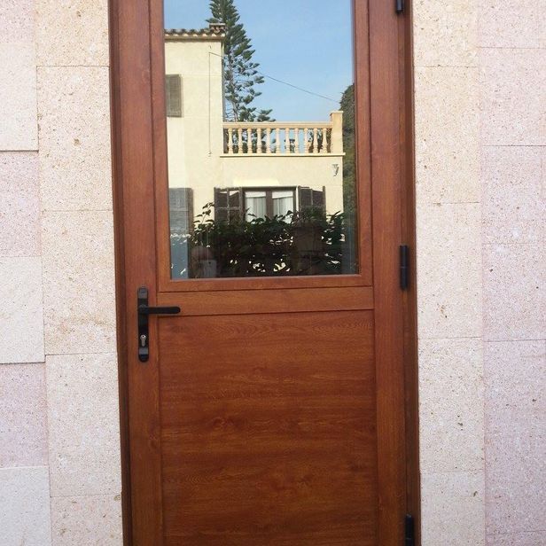 Puerta de madera marrón con panel de vidrio que refleja un edificio, situado en una pared de piedra.