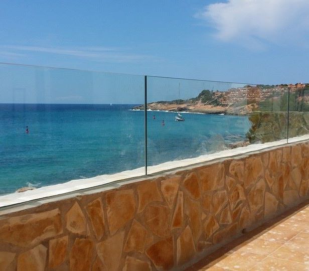 Vista al mar desde un balcón de piedra y cristal. Agua azul, cielo despejado, un barco y un acantilado a lo lejos.