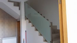 Escalera con barandilla de cristal, escalones grises y paredes blancas.