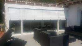 Patio exterior con persianas blancas, ventanas de vidrio y muebles negros.